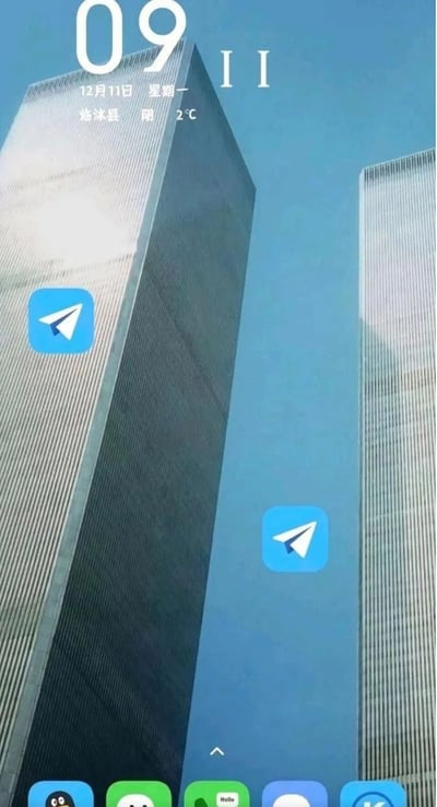 图片展示了一张智能手机的主屏幕截图，背景为两座高耸的摩天大楼（外观类似双子塔）。屏幕顶部显示时间为“09:11”，日期“12月11日 星期一”，地点“光沐县”，天气“阴”，气温“2°C”。屏幕左侧和右侧中间位置各有一个蓝色纸飞机图标的Telegram应用图标，底部dock栏包含QQ、微信、“Hello”及另一个应用图标。画面中时间“09:11”与双子塔背景结合，可能存在对特定历史事件的暗示，具有一定争议性。