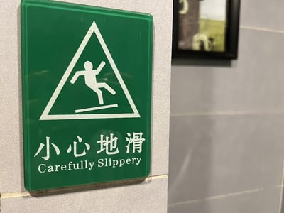 这是一张常见的警示标志照片，绿色三角形内有小人滑倒的图案，下方配有中文和英文警告文字。中文为“小心地滑”，而英文翻译成了“Carefully Slippery”，语法不通顺，给人带来幽默感，经常出现在网络上被调侃为“中式英语”或“中式翻译”笑料。这种翻译失误，形成了谐音梗或语法梗，成为网友互相调侃的对象。

双关/谐音/暗示说明：
英文“Carefully Slippery”其实应该是“Caution: Slippery Floor”或“Be Careful: Slippery”，现有翻译直接把单词照搬，制造了英语语法上的幽默效果。
