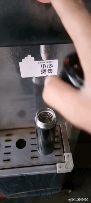 这是一段竖屏动态视频，展示了在饮水机热水出口下，一个金属易拉罐接热水时，随着热水的持续冲刷，易拉罐逐渐“变矮”，最后只剩下罐口部分，制造出一种热水把易拉罐“冲没了”的搞笑视觉效果，实际上是通过移动易拉罐和调整拍摄角度制造的错觉，配上饮水机“小心烫伤”的提示，反差感拉满，十分滑稽。