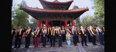 图片展示了一群人在传统中式建筑前的集体活动场景。前景中，数十位成年人身着深色西装，颈系黄色围巾，整齐站立在台阶上；人群中间有一位身着蓝色传统服饰的儿童。背景是一座具有典型中国古建筑特征的 pavilion，其拥有双层飞檐、红色立柱和雕花装饰的木质结构，屋顶覆盖着红色瓦片，周围环绕着茂密的绿树，整体氛围庄重，推测可能是一场与传统文化相关的仪式或纪念活动。