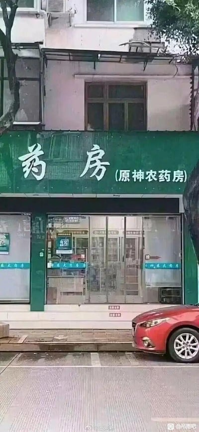 这是一张街景实拍图，画面主体是一家临街的药房店铺。店铺的绿色招牌上用醒目的白色大字标注“药房”，右侧括号内写着“(原神农药房)”，实际上该店铺原名为“神农农药房”，简称后拆分的文字巧合与热门游戏《原神》以及“农药”形成关联，产生了趣味玩梗的效果。店铺采用玻璃门橱窗设计，透过玻璃能看到内部摆放的货架，玻璃门下方的蓝色字样标注着“神农大药房”。店铺门前的人行道地面湿润，看起来刚下过雨，一辆红色轿车停在店铺门前的路边，背景是带有窗户和空调外机的居民楼墙面，整体是日常的城市街景。

这个招牌的趣味点在于名称的巧合：原“神农农药房”去掉“农”字的偏旁后，被拆分为“原神”+“农药房”，和热门游戏《原神》以及常见的“农药”（如农用农药、游戏《王者荣耀》的俗称）形成关联，引发网友的趣味联想。