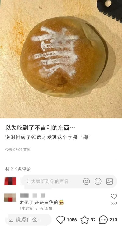 这是一张社交平台分享截图，上半部分是实拍的放在木质砧板上的棕色圆形面包，面包表面用白色粉末写了一个汉字，正放状态下字形非常像寓意不吉利的“奠”字；下半部分是发布者的配文，称自己一开始看到这个字以为买到了不吉利的食物，直到将面包逆时针旋转90度之后，才发现这个字实际是“椰”，代表这是椰馅面包。截图下方还展示了其他网友的评论“太像了 还是白色的”，以及点赞1086、收藏32、评论219的互动数据，整体是因文字摆放角度问题看错文字产生的搞笑乌龙内容。