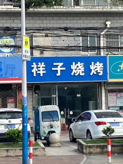 这是一张街头实拍的横图，展示了居民区楼下的「祥子烧烤」店铺：蓝色招牌配醒目白色大字，店铺门前的通道里停着一辆前脸有破损的电动三轮车，两侧还停着两辆白色轿车；画面左侧有一个「校车停靠站点」的交通标识，背景是灰色砖石外墙的居民楼，空中布满交错杂乱的电线，整体是充满生活气息的日常城市街景，没有谐音梗或双关内容。