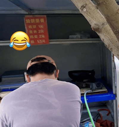 这是一张拍摄于街头烧饼摊的照片。图片中央是一名戴着棒球帽的男子背影，似乎正在摊位内准备食材。摊位上方悬挂着一块红色的价目表，写有“烧饼里脊”，但价格设置有趣，几乎所有主食的价格都一样：烧饼2元，里脊2元，鸡蛋2元，但烧饼单独只需1元。这种设定让人觉得有点“物价逻辑混乱”，引人发笑。图片左上角还加了一个大哭笑表情，更加突出搞笑和无厘头氛围。

此图有明显的网络幽默、反差、无厘头风格，没有明确谐音梗或明显暗示，但价格表的设置本身形成了一种“算不过来的梗”。