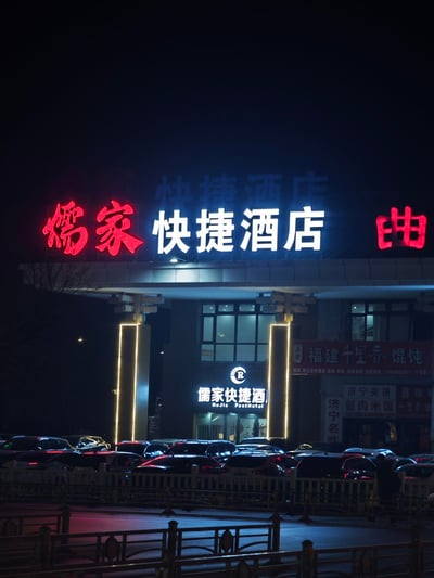 夜晚拍摄的儒家快捷酒店外观，酒店招牌以红色和蓝色霓虹灯醒目点亮，左侧为红色"儒家"二字，中间为蓝色"快捷酒店"四字，右侧部分可见红色"由"字。酒店入口处有两根立柱装饰着黄色灯带，下方停车场停有多辆汽车。周围还能看到"福建千里香馄饨"、"济宁夹饼"、"济宁甏肉米饭"等商铺招牌，整体呈现出城市夜间的商业氛围。
