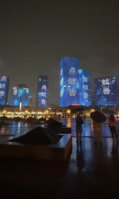 画面为夜晚城市广场，多个高楼外墙打出蓝色大字“点蚊香”，在灯光和雨后地面反光的映衬下格外醒目。地面有几位打伞路人在观望。这种将日常生活中常见的防蚊建议做成巨型LED灯光秀，形成强烈视觉反差与幽默效果。图片因此在网络流行，表现了现代都市人的无奈与幽默感。

双关/暗示分析：
“点蚊香”一般指生活小常识，这里夸张展示，形成人与城市生活的荒诞反差。