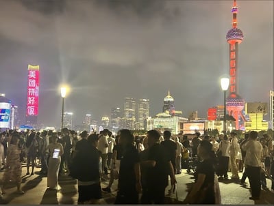 这张图片拍摄于上海外滩的夜晚，可以看到大量市民和游客聚集在江边，人头攒动，气氛非常热闹。远处是著名的东方明珠塔，亮眼的灯光让整个城市夜景显得格外璀璨。左侧高楼上巨大的『美团拼好饭』广告牌极为醒目，在现代都市灯光和人流的对比下，营造出一种都市快节奏与互联网生活的既视感。

不少人使用手机拍照或录视频，展现了人们夜生活的丰富样貌。整体气氛略带都市的『挤』或『卷』感，也暗合当今城市人群的娱乐、消费与社交现状。广告词“拼好饭”有种接地气的调侃意味，暗示大家都在拼团、精打细算、集体消费的大环境中。

没有明显的双关或谐音梗，但现代都市感、互联网广告包围氛围非常突出。