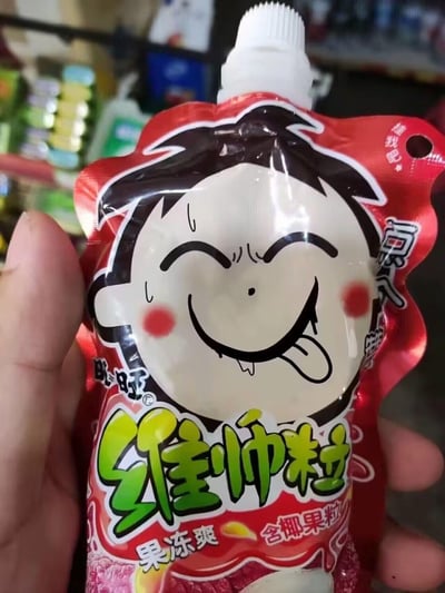 这是一只手拿着旺旺维多粒果冻爽的吸吸袋包装，包装主色调为红色，印着魔性表情的旺仔卡通形象：眯起双眼，吐着舌头，嘴角流出口水，脸上还挂着汗珠，脸颊泛红，表情滑稽又满足，看起来十分搞笑。包装标注该产品为含椰果粒的果冻爽，是旺旺旗下的零食产品。