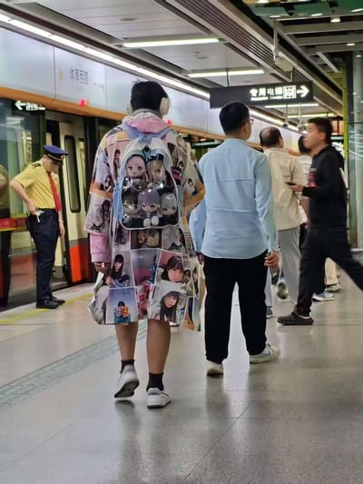 地铁平台场景中，一名乘客从背后看穿着印有大量动漫人物图像的拼贴风格外套，背着透明背包，内装多个动漫角色毛绒玩偶。周围有其他乘客行走或站立，包括一名穿浅蓝色衬衫的男子和一名使用手机的黑衣男子。背景可见地铁列车门，一名穿黄色制服的安全人员站在车门旁，上方有“体育西路”和“专用电梯”方向指示牌。整体展现了城市地铁中年轻人独特的二次元时尚风格与日常通勤场景的融合。