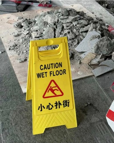 图片中展示了一个黄色的湿滑地面警示牌，放置在施工场地的碎石堆前。警示牌上方用英文写着"CAUTION WET FLOOR"，下方配有中文"小心扑街"的醒目提示，中间是一个红色的滑倒人形警示图案。背景中可以看到散落的混凝土碎块和施工材料，显示出这是一个正在进行维修或建设的区域。这种将正式警示语与口语化中文结合的方式，形成了一种幽默效果。