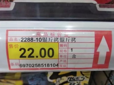 这是一张超市货架的实拍图，核心内容是一张红白色的商品标价签，标签上的品名栏错误打印成了“2288-10锟斤拷锟斤拷”，“锟斤拷”是中文在不同字符编码转换出错时会出现的典型乱码文本，属于互联网程序员圈层的常见梗；标签同时标注该商品售价22.00元，规格为1盒，商品条码为6970258518104，标价签右侧带有红底白色向上箭头用于指示商品摆放位置，标签下方可以看到模糊的商品包装局部。