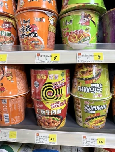 这是一张实拍超市货架的照片，上层货架摆放了康师傅品牌不同口味的桶装泡面，价格标签显示单价均为5元；下层货架主要摆放统一品牌的不同口味桶装泡面，单价标注为4.5元，有趣的是一罐榄菊品牌的桶装蚊香被错误摆放在下层泡面区域，放在统一红烧牛肉泡面桶上方，错放在泡面商品堆里，形成了强烈的反差感，制造出很诙谐的效果，仿佛蚊香成了泡面品类的商品，非常巧合搞笑。