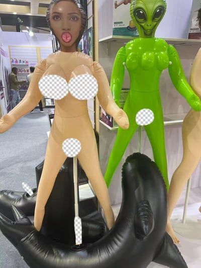 这张图片展示了在一个展览或贸易展上展出的充气娃娃。前景中有两个主要的充气玩偶：一个是米色的人形玩偶，有着夸张的惊讶表情，嘴巴张开；另一个是绿色的外星人形象玩偶，有着大大的眼睛和典型的外星人特征。两个玩偶的隐私部位都用透明的方格图案进行了模糊处理。背景中可以看到展览的其他部分，有货架、产品和几个人在参观。