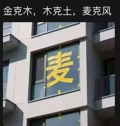 一栋建筑的窗户上用黄色胶带贴着巨大的“麦”字，上方配有文字“金克木，木克土，麦克风”。图片通过语义梗制造笑点，将传统五行相克理论中的“金克木，木克土”句式，无厘头地接续为“麦克风”，形成幽默的反差效果。