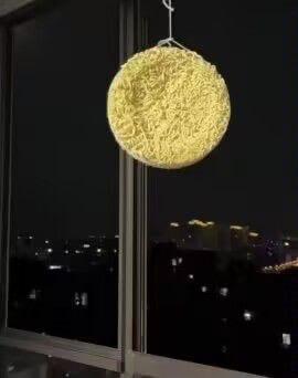 夜晚的室内窗边，一个圆形的泡面被悬挂在窗户内侧，借助窗外漆黑的夜景背景，从特定角度看去，泡面的形态和光线反射效果让它看起来像是夜空中的月亮，形成了有趣的视觉错位效果，充满了搞笑的反差感，是网友的创意整活内容。