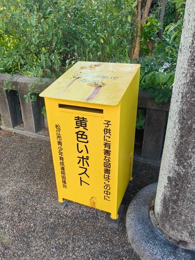 这是一张户外实拍照片，主体是一个亮黄色的立式公共回收箱，箱体标注为“黄色邮箱”，由松江市青少年育成联络协议会设置，专门用于投放对儿童有害的图书。箱子顶部印有“请勿放入垃圾”的提示，箱体有轻微使用污渍，放置在铺有碎石的地面上，背景是茂密的绿植与石质护栏，属于户外公共区域的公益设施场景，无谐音梗或双关含义。