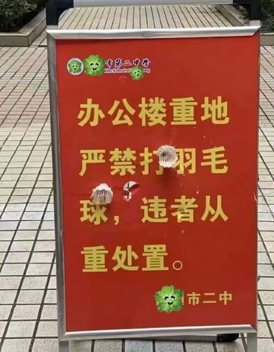图片中展示了一块红色的警示牌，位于市第二中学的办公楼区域，放置在铺有方形地砖的地面上。警示牌主体为红色，上方印有“市第二中学”字样及绿色卷心菜形状的笑脸贴纸，中间用黄色大字写着“办公楼重地 严禁打羽毛球，违者从重处置。” 有趣的是，警示牌上有明显的破损痕迹，其中“打”字和“球”字附近各有一个孔洞，并有一个白色的羽毛球被卡在“打”字上方，形成了“禁止打羽毛球”的标语被羽毛球“命中”的讽刺画面，充满了校园生活的幽默感。