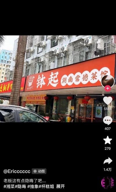 这是一张竖屏的短视频平台截图，画面主体是街边的一家湖南常德菜馆，店铺的橙红色招牌上印有白色的“钵起 湖南常德菜馆”字样与餐饮类logo，菜馆门口挂有红灯笼，门口坐着一名人员，近处有车辆和行道树，背景是安装了防盗网和空调外机的居民楼。画面右侧是短视频平台的交互按钮，分别显示点赞5184、评论407、收藏279、转发1.4万。发布者配文调侃店名隐晦，该店名“钵起”读音与中文不雅词汇发音相近，因此引发网友讨论，觉得取名十分抽象有梗。