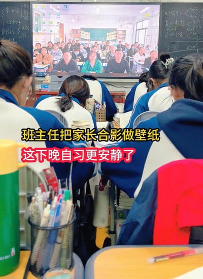 这是一张晚自习教室的实拍照片，画面中穿着蓝白相间校服的学生们坐在课桌前，前方的大屏幕上显示着全班家长的合影，被班主任设置成了屏幕壁纸。图中叠加了文字：“班主任把家长合影做壁纸 这下晚自习更安静了”，幽默地展现了这个巧妙的班级管理方法——通过让家长“在线盯着”的心理暗示，让学生们在晚自习时更加自觉安静，充满了校园生活的趣味感，没有双关或谐音梗，仅利用心理效应来维持自习纪律。