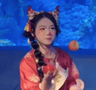 一位身着红色传统服饰、头戴装饰性发冠的女子，在蓝色背景前与一颗红色果实（疑似桃子或苹果）进行动态互动。她手持果实，通过手部动作和表情变化展现出专注且略带俏皮的状态，果实随着她的动作呈现出自然的动态轨迹，整体画面生动有趣，充满传统与现代结合的创意感。