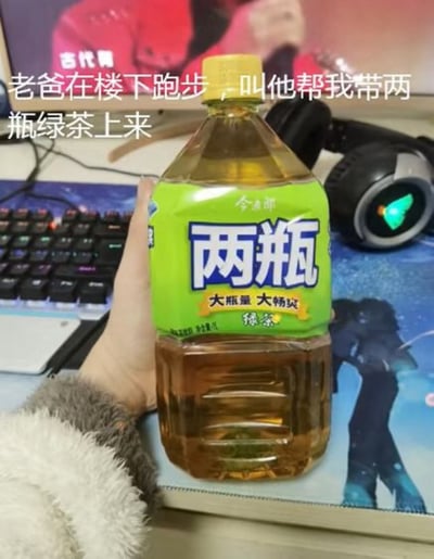 图中展示一只手拿着一瓶今麦郎绿茶，瓶身标签醒目地印着“两瓶”大字，旁边标注“大量 大畅饮”。背景可见电脑屏幕、键盘和耳机，营造出居家场景。图片上方配有文字：“老爸在楼下跑步，叫他帮我带两瓶绿茶上来”，幽默之处在于“两瓶”并非指两瓶独立包装，而是这瓶名为“两瓶”的大容量绿茶，形成因文字歧义产生的搞笑误会。