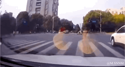 这是一段行车记录仪拍摄的动态画面：在城市路口等待红灯时，画面右侧的白色SUV正常通行，前方路口可见其他车辆与行人。突然一只浅灰色运动鞋从画面外飞来，重重砸在拍摄车辆的前挡风玻璃上，还被启动的雨刮器扫动了一下，整个过程意外又搞笑，让人猝不及防。
无双关、谐音梗或暗示内容。