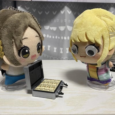 图中是《LoveLive!Sunshine!!》的渡边曜与小原鞠莉的毛绒玩偶，渡边曜玩偶旁摆放着一个打开的、装满“钞票”的手提箱，小原鞠莉玩偶被加装了夸张的凸眼特效配件，呈现出看到巨款后极度震惊的神态，是ACG爱好者用玩偶搭建的趣味“交易现场”摆拍场景，充满搞笑感。