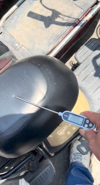 这是一张实拍竖图，展示了夏季户外场景下，拍摄者手持探针式温度计测量停放在室外的电动车黑色坐垫温度的画面，温度计显示数值达到88.6摄氏度，画面还露出了电动车的脚踏、车架结构以及拍摄者的腿脚部分，非常直观地体现了夏日烈日暴晒后户外物品温度极高的酷暑状态，冲击力很强，很容易让大众联想到夏天坐烫座椅的糟糕体验。