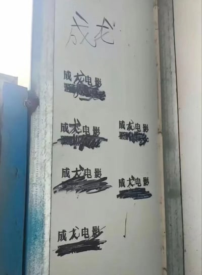 图片展示了一面白色墙壁，墙面上有多处手写的黑色涂鸦文字。最上方用较大字体写着“成龙”二字，其下方分布着多个“成龙电影”字样的涂鸦，部分涂鸦存在涂抹、划掉或字迹模糊的情况。墙壁左侧可见一扇蓝色的门，整体场景像是在室外的公共区域，如学校、社区或地下通道等。