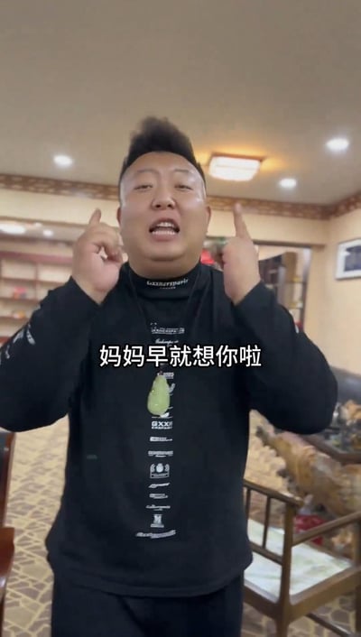 图中男子身着黑色带有白色图案的毛衣，胸前佩戴绿色吊坠，站在铺有花纹地毯的室内场景中，背景可见书架、木质椅子和装饰摆件。他双手食指指向上方，表情真挚，配合文字'妈妈早就想你啦'，整体传递出家人间温暖的思念情感。