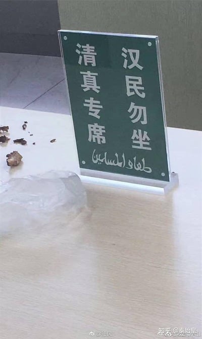 照片显示餐厅或食堂桌子上的标识牌，上面写着“清真专席 汉民勿坐”，并配有阿拉伯文。该牌旨在为清真饮食顾客设置专用区域，但“汉民勿坐”的用词显得争议十足，容易引发网友对民族之间隔离或误解的吐槽。桌面有塑料袋和残渣，环境很普遍。这类梗图常被网友拿来调侃隔离或餐饮歧视现象。

无明显双关或谐音，但民族隔离暗示常作为讨论点。