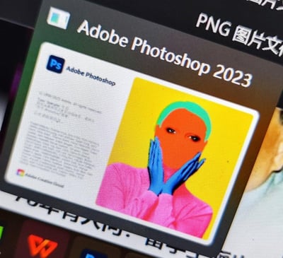 这是一张电脑屏幕的截图，展示了Adobe Photoshop 2023的启动界面。界面右侧是一幅具有波普艺术风格的高饱和色彩人像作品：人物有着青绿色头发，脸部为橙色，双手是蓝色，身着粉色上衣，双手放在脸颊两侧，背景为明亮的黄色；界面左侧是软件相关的文本信息区域，包含软件名称等内容，左下角还有Adobe Creative Cloud的标识。整体视觉风格鲜明前卫，充满艺术感。
无双关、谐音梗或暗示内容。