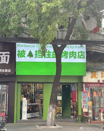 这是一张街头店铺的实拍静态图，画面中心是一家绿色门头的烤肉店，一棵大树恰好生长在店铺正前方，遮挡住了招牌的部分区域。店主将这一不利情况转化为店铺特色，设计了趣味招牌：中文为“被🌲挡住的烤肉店”，用圣诞树图标替代“树”字，上方搭配英文“Barbecue restaurant blocked by trees.”，充满幽默感。店铺左侧是带有“面 民生路店”字样的面馆，右侧是“夏氏”相关店铺，烤肉店内部墙面贴满了各类人物影视海报，门口摆放着小桌椅，整体呈现出充满生活气息的街头场景。