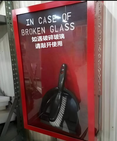 图片展示了一个红色边框的玻璃应急箱，箱内放置着黑色的簸箕和刷子。箱子正面有中英文双语提示：英文写着"IN CASE OF BROKEN GLASS"，中文翻译为"如遇破碎玻璃 请敲开使用"。这个提示存在明显的逻辑矛盾和翻译问题，因为遇到破碎玻璃时需要敲碎这个玻璃箱才能取出清洁工具，形成了"为了清理碎玻璃而制造更多碎玻璃"的荒诞场景。
