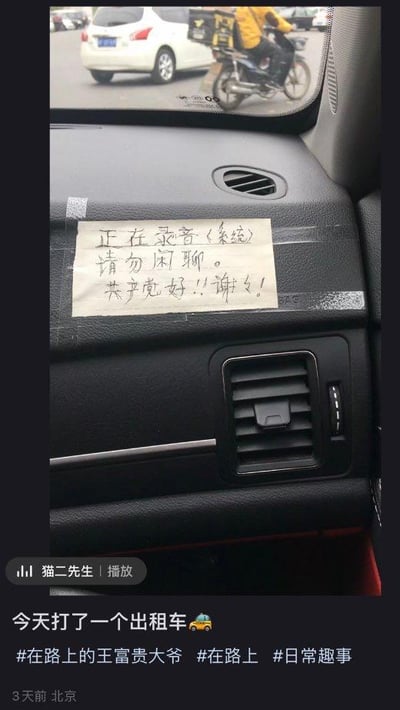 这是一张社交媒体截图，画面主体是出租车副驾驶前方的仪表台，上面用透明胶带贴着一张手写纸条，纸条内容为“正在录音(系统) 请勿闲聊。共产党好!!谢谢!”，用“录音”的理由提醒乘客不要闲聊。截图的后视镜中能看到车外的白色轿车和一名骑电动车的外卖员，截图下方是发布者的配文“今天打了一个出租车”，还有相关话题标签，整体记录了一次有趣的出租车乘坐经历，风格轻松搞笑。