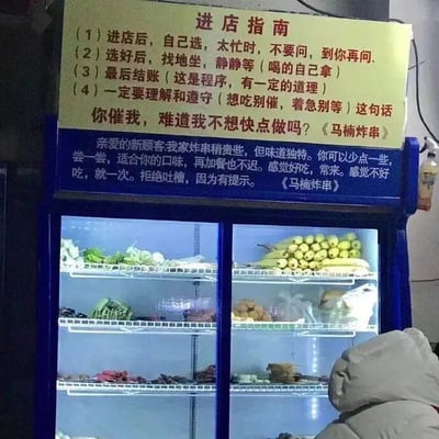 这是一张线下小吃店的实拍静态图，画面主体分为上下两部分：上方是一块黄底红字的“进店指南”提示牌，内容直白又硬核，明确告知顾客进店后的流程：自行选餐、选好就坐等待、最后结账，还着重提醒顾客在店家忙碌时不要询问、不要催促，甚至直接表示“着急别等”；提示牌下方还有一段给新顾客的说明，提到店铺炸串价格稍高但味道独特，建议顾客先少量尝试，合口味再加餐，同时拒绝顾客吐槽。提示牌的两处位置都标注了店名“马桶炸串”。画面下方是一个蓝色边框的玻璃冷藏展示柜，内部分层摆放着各类待炸的蔬菜、豆制品等食材，右下角有一位穿着浅灰色连帽厚外套的顾客，背对着镜头，似乎正在挑选柜内的食材。整个画面充满了这家小吃店随性、直白的经营风格。