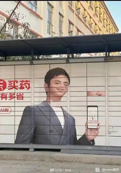 这是一张设置在智能快递柜柜面的购药平台广告海报，因其中一个快递柜门被打开，导致海报画面被柜门分割错位：原本正常的男性广告模特脸部被柜门切割，呈现出怪异滑稽的歪脸效果，模特手持展示购药APP的手部也和身体部分分割开来，广告上的文字“买药有多省”“分购”以及手机屏幕上的“打开购药神器 低价放心买药”也被柜门打乱布局，原本正经的商业广告意外变成了充满荒诞感的搞笑画面，视觉反差强烈，毫无刻意创作的双关或谐音梗，完全是场景错位带来的意外笑点。
