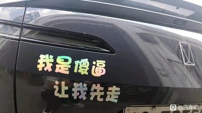 这是一张实拍的汽车局部照片，黑色汽车的车身侧面贴有镭射反光效果的趣味车贴，车贴文字为“我是傻逼 让我先走”，采用自黑自嘲的幽默话术，试图以此方式请求其他道路车辆为自己让行，风格接地气又搞笑，用夸张的自嘲来制造喜剧效果。
