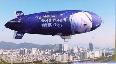 这是一张在晴朗天气下拍摄的户外照片，画面中一艘深蓝色星空底色的宣传飞艇漂浮在城市上空，飞艇侧面印有手游《胜利女神：妮姬》的白发红瞳角色形象，以及韩文宣传语“가장 아름다운 승리를 안겨줄게”（中文意为“会为你带来最美丽的胜利”），还有“NIKKE 2ND ANNIVERSARY”的周年庆标识，尾翼处也印有“NIKKE”的游戏品牌字样。背景是密集的城市高层建筑群与远处的山峦，整体场景醒目吸睛，是游戏周年庆的线下空中宣传形式。