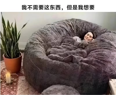 图片展示了一个人舒适地躺在一个巨大的灰色豆袋椅中，豆袋椅大到看起来像一个可以让人完全陷进去的巢穴。人物面带满足的微笑，显得非常放松。图片上方配有文字'我不需要这东西，但是我想要'，幽默地表达了人们对非必需品的强烈渴望心理。
