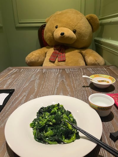 一只戴着红色格子领结的巨型棕色泰迪熊正端坐在餐桌前，面前摆放着一盘清炒绿叶蔬菜，旁边配有带花纹的小碗（内有白色勺子）和一杯白水。餐桌为木质纹理，背景是浅绿色墙面与白色装饰线条，营造出温馨的餐厅或家庭用餐氛围。泰迪熊的可爱造型与日常食物形成有趣对比，显得既治愈又富有生活气息。