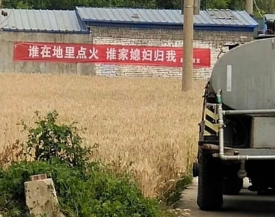 图片呈现了乡村田间的场景：前景是路边的绿植，中间是一片金黄的成熟麦田，麦田旁的砖墙上悬挂着一条醒目的红色标语，写着“谁在地里点火 谁家媳妇归我”，画面右侧停着一辆洒水车。这条标语用夸张戏谑的玩笑式表述，来宣传禁止田间焚烧秸秆的规定，用极具冲击力的搞笑内容来强化警示效果，没有双关、谐音梗等设计，纯靠夸张的“惩罚”设定吸引注意。