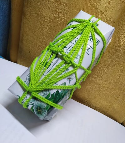 图片中展示了一个被亮绿色毛线紧密缠绕的白色牛奶盒，毛线在盒身形成交叉编织的网状结构，顶部打有结固定。牛奶盒表面印有绿色文字和图案（部分可见“生活”“证”等字样及植物图案），放置在白色平面上，背景为带有纹理的金色布料，整体呈现出一种创意手工改造的温馨感，可能是家居装饰或收纳小技巧的展示。