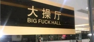 这是一张室内拍摄的场所标识牌照片，标识牌为黑底配金色边框，上方是简体中文“大操厅”，下方的英文翻译却错误使用了带有低俗冒犯性的词汇“FUCK”，写成“BIG FUCK HALL”。“大操厅”原本指用于操练、集合的正式场所，正确的英文翻译不应使用该词汇，这个低级的翻译失误让原本正式的标识牌产生了荒诞搞笑的反差效果。背景中可以看到模糊的室内环境和一个人物的身影。