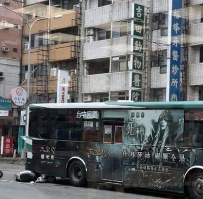 这是一张拍摄于台湾台中的街头照片，画面中一辆台中客运的公交车车身印有游戏《天堂M》的宣传广告，广告内容为「化身死神 碾压全场」，并配有表情冷峻的死神形象。公交车旁的地面上有一人倒地，从拍摄角度看仿佛是被广告中的「死神」「碾压」，形成了强烈的视觉错位效果，营造出戏剧性的趣味感。背景是典型的台湾城市街景建筑，可见「吉田动物医院」「裕峰牙医诊所」等店铺招牌，整体场景是真实拍摄的巧合，并非刻意的事故或P图改编。