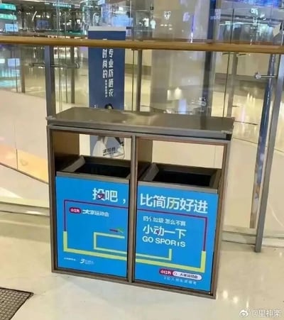 这是一张拍摄于商场公共区域的实拍图，主体是一个双桶式公共垃圾桶，桶身贴有小红书“大家运动会”主题的趣味宣传标语。左侧桶身印有“投吧，”，右侧桶身印有“比简历好进 扔个垃圾怎么不算 小动一下 GO SPOR1S”，其中“SPOR1S”是“SPORTS”的谐音拼写。标语巧妙结合当下年轻人求职难、简历投递后入职成功率低的职场痛点，将扔垃圾这个简单的日常小动作与求职入职对比，用自嘲的方式制造趣味笑点，同时贴合“大家运动会”倡导日常随时随地做小运动的活动理念。背景可见商场的玻璃护栏与其他商业标识，图片右下角带有微博用户@里神楽的水印。