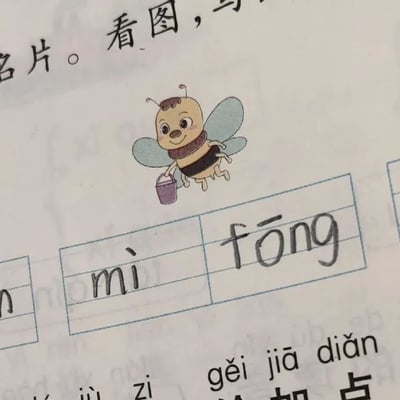 这是一张儿童作业本页面的特写照片，画面中央是一只卡通蜜蜂插图：蜜蜂有着圆脸蛋、微笑表情，身体呈黄黑条纹状，翅膀为浅蓝色，正手持一个紫色小桶飞行。插图下方是拼音练习格，手写着“mì”和“fōng”（推测应为“fēng”，可能是书写错误）。页面周围可见部分印刷文字，包括“看图，写...”（提示学生看图写字）和“gěi jiā diǎn”（给加点字，可能指注音或标调）。整体风格充满童趣，适合低龄儿童学习使用。