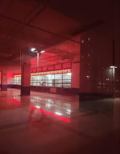 这是一张深夜在空旷室内大厅拍摄的照片，整个空间被红色灯光笼罩，光洁的地面清晰反射出灯光与标语的倒影。大厅的玻璃窗上悬挂着红色LED滚动标语，无休无止地重复显示「你好你好你好你好你好你好你好你好你好你好你好你好你！」，远处的区域也有一处相同的标语。场景中没有人物，空旷的环境加上重复的标语，营造出一种既规整又带着荒诞诡异感的氛围。