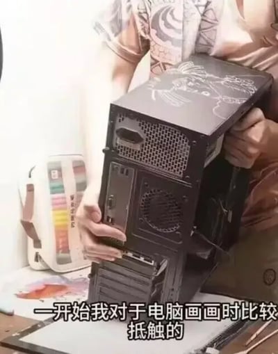 这是一张实拍静态图，画面中一名穿着带有龙纹印花短袖的人双手捧着一台黑色电脑机箱，机箱顶部有手绘的图案，背景桌面上摆放着彩色马克笔收纳盒和画纸类的绘画工具，图片底部配文“一开始我对于电脑画画时比较抵触的”，用在实体电脑机箱上作画的方式玩梗，调侃部分手绘创作者对数字绘画存在抵触、偏好传统手绘创作的心态。