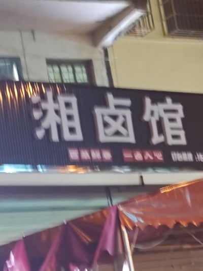 这是一张实拍的街边卤味餐馆招牌照片，黑色竖纹背景上是醒目的白色大字“湘卤馆”，下方配有红色小字标语“百味鲜醇 一卤入心”，右侧还有部分被遮挡的联系电话。招牌下方可见紫红色的遮阳篷，背景是带有窗户和防护栏的居民楼墙面，呈现出典型的街边社区小店的场景，图中没有双关、谐音梗或暗示内容。